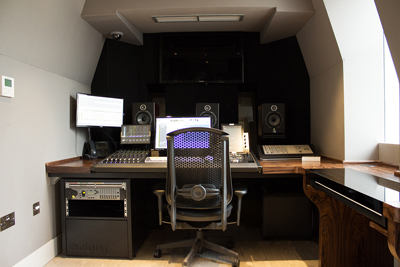 A music production desk with computer monitors, studio monitors, a mixing console, and an ergonomic chair.