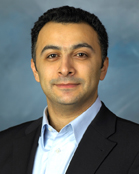 A man with dark hair and tan skin wearing a light blue shirt and black blazer against a blue background.