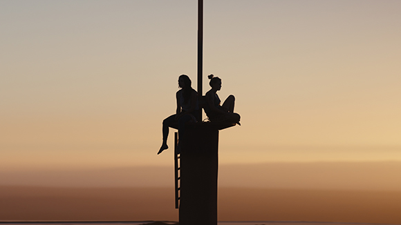 Silhouettes of two people sitting atop a tall pole against a sunset sky.