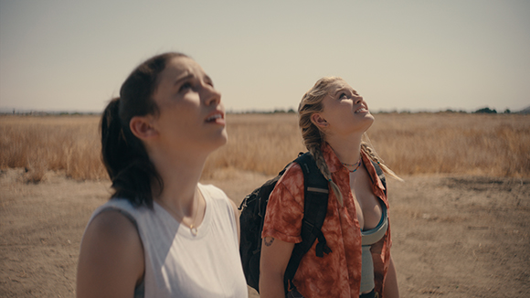 Two young women standing in a dry field, looking upward toward the sky with curious expressions.