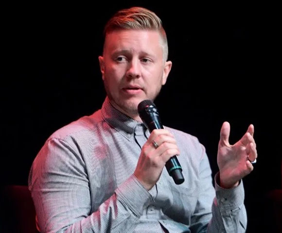 A man in a grey striped shirt speaks into a black microphone while gesturing with his left hand.