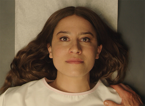 A woman with long wavy brown hair lies on a white surface, looking directly forward.