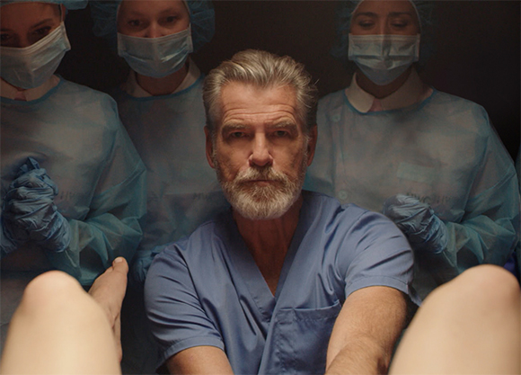 A man with grey hair and a beard in blue scrubs sits before three masked medical workers in blue surgical attire.