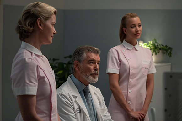 A man in a white lab coat sits while two women in pink nursing uniforms stand beside him.