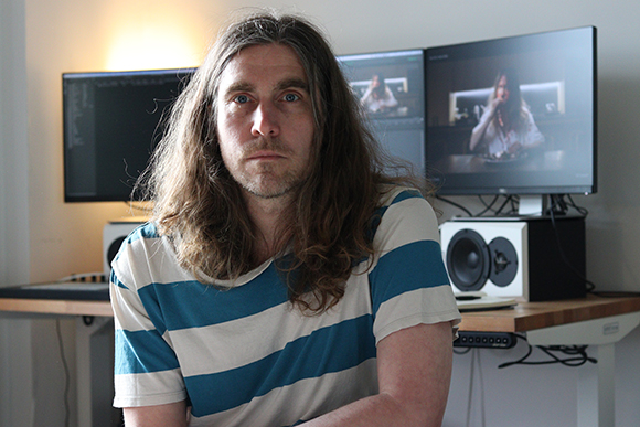A man with long brown hair wearing a striped shirt sits in front of two computer monitors and studio speakers.