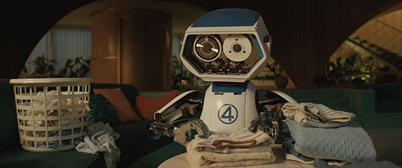 A white and blue robot stands behind a pile of folded laundry in a dimly lit room.