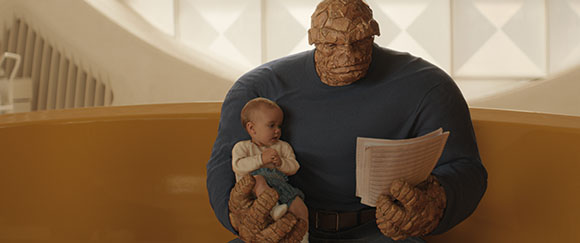 A large, orange, rock-skinned humanoid sits holding a small baby and a sheet of paper.