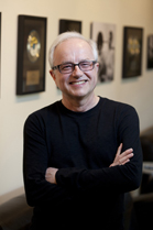 A smiling man with glasses and a black long-sleeved shirt stands with his arms crossed in front of framed pictures.