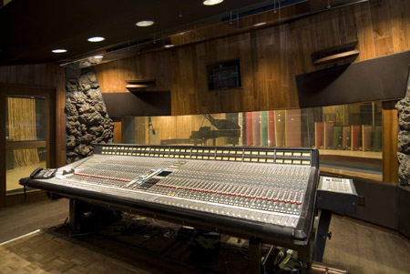A large audio mixing console sits in a wood-paneled recording studio with a view into a live room.