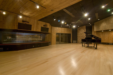 A spacious music studio with light wood floors, wood-paneled walls, and a black grand piano.
