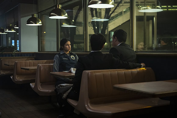 A female police officer sits in a diner booth talking to two men in suits.