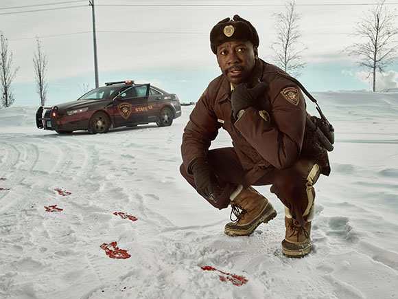 A man in a brown uniform crouches in the snow near red stains and a state trooper car.