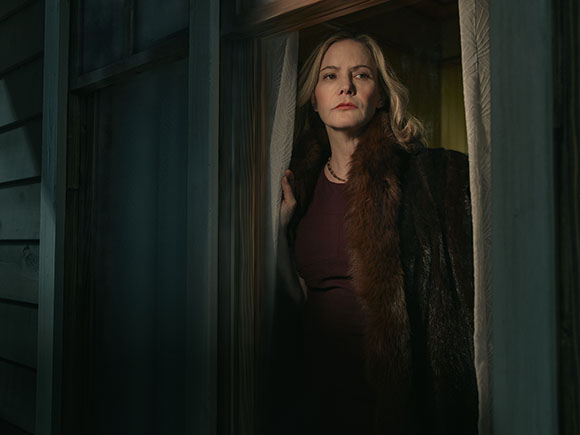 A woman wearing a dark top and a brown fur coat looks out from a doorway.