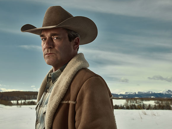 A man wearing a brown cowboy hat and a tan shearling jacket stands in a snowy landscape.