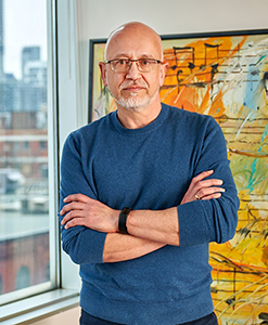 A bald man with glasses and a goatee wearing a blue sweater stands with arms crossed in front of artwork.