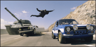 A man flies through the air between a tank and two cars driving on a highway near a rocky hill.