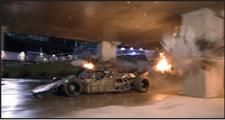 A low-profile racing car with an exposed metal frame undergoes an explosion under a concrete structure.