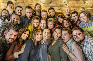 A large, diverse group of smiling adults posing closely together for a group photo against a wooden wall.