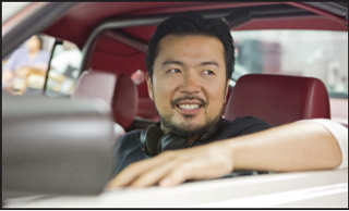 A man with dark hair and a beard sits in the back of a car with red leather seats.