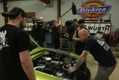 A film crew with cameras and a boom mic records a man working on a lime green car engine in a garage.