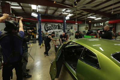Film crew filming people working on a green classic car inside a large automotive garage.