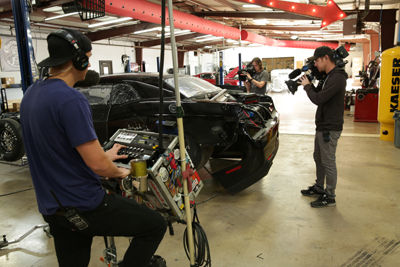A camera operator and sound technician film a black race car inside a professional garage.