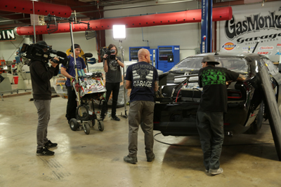 A film crew with cameras and lights filming a black muscle car inside a large automotive garage.
