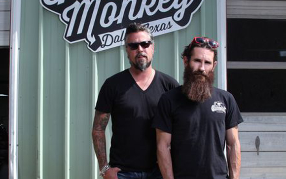 Two men with facial hair standing in front of a sign that reads Monkey Dallas Texas.