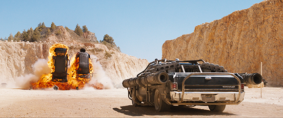 A modified black car in a desert quarry as a person on a flaming hoverboard flies into the distance.