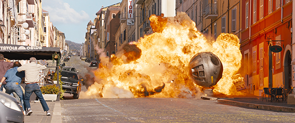 A massive orange fireball and debris erupt in a narrow European street near two people.
