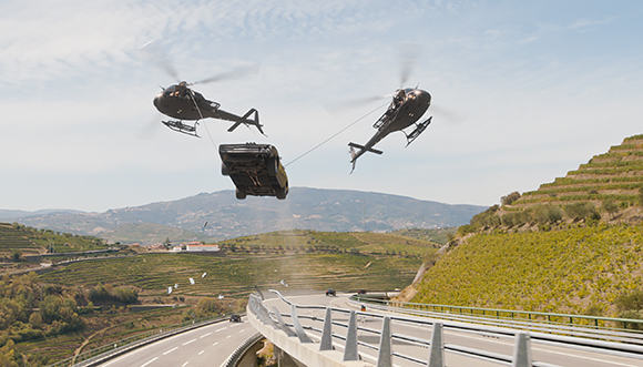 Two helicopters use cables to lift a black car over a winding highway in a hilly landscape.