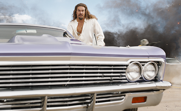 A man with long hair and a white shirt stands behind a purple vintage car amidst smoke.