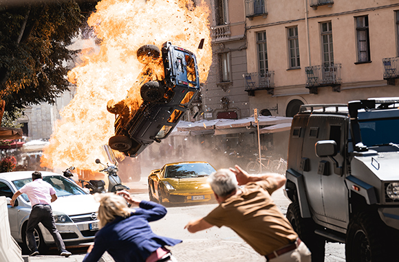 A dark SUV flips through a massive orange fireball in a city street as people react in the foreground.