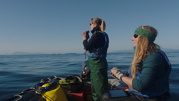 Two women in life vests stand on a boat looking out over a calm blue ocean under a clear sky.