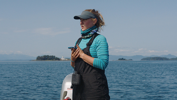 A woman wearing a sun visor and life vest stands on a boat against a backdrop of blue ocean and distant islands.