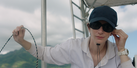 A person in a white shirt, dark cap, and sunglasses holds a thin rope against a cloudy sky and green hills.