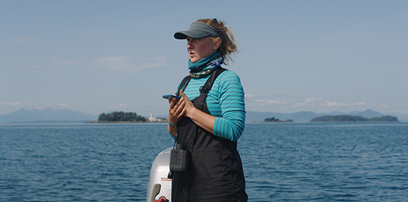 A woman wearing a visor, blue striped shirt, and black overalls stands on a boat overlooking the ocean.