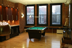 A pool table sits in a room with exposed brick walls, wooden floors, and large windows overlooking a city.