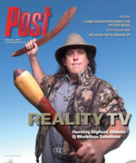 Man in hat and camouflage jacket holds two wooden weapons, one curved and one club-shaped, outdoors.