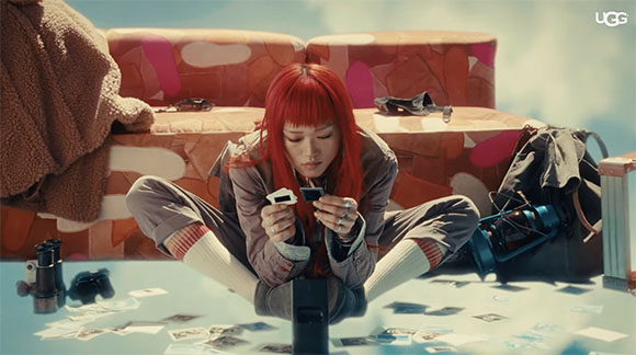 Person with bright red hair sits on the floor surrounded by photos and vintage cameras.