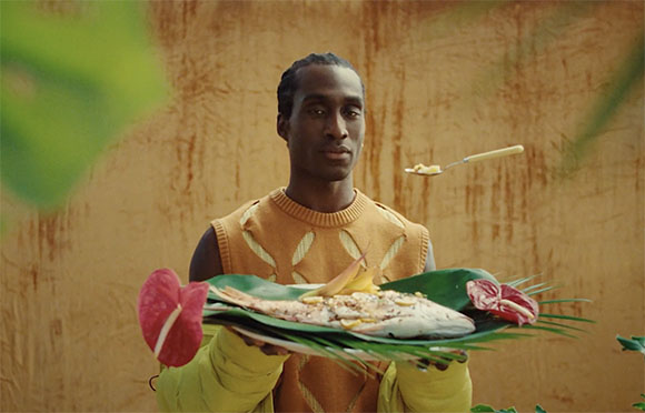 A man in an ochre sleeveless knit top holds a large green leaf platter with a whole cooked fish.