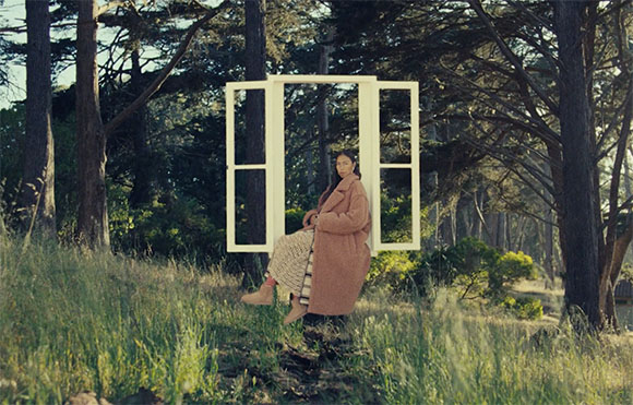 A person in a pink coat sits within a white window frame standing in a sunlit forest.