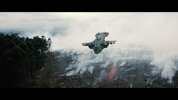 A dark, metallic spacecraft flies over a charred, smoky landscape of cleared forest.