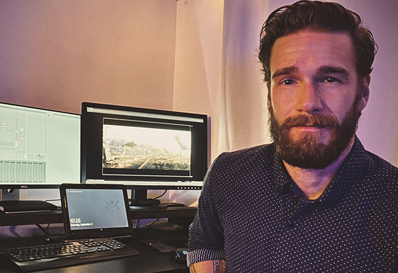 A bearded man in a patterned shirt sits in front of two computer monitors in a dimly lit room.
