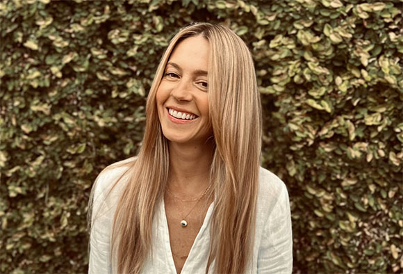 A smiling woman with long blonde hair wearing a white shirt against a dense green leaf background.