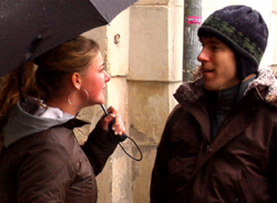 A woman holding a black umbrella talks to a man wearing a dark beanie and winter jacket.