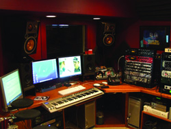 A music studio desk with dual monitors, a MIDI keyboard, studio monitors, and various audio equipment.