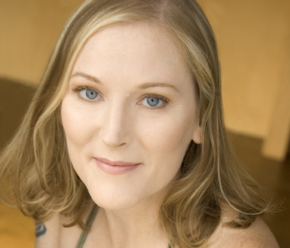 A close-up portrait of a woman with blonde hair and blue eyes looking directly at the camera.