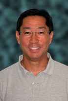 A smiling man with dark hair and glasses wearing a grey polo shirt against a dark blue background.