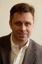 A man with short brown hair wearing a white collared shirt and a brown blazer.